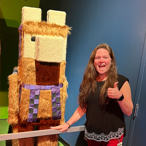 Woman with long hair giving a thumbs-up signal while standing next to a life sized Llama mob sculpture.
